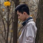 Side view of a young man with headphones, leaning on a tree in an autumn forest.